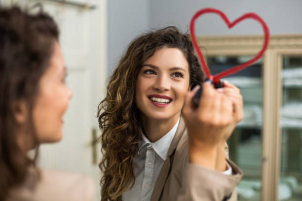 Woman smiling at her reflection in a mirror, holding a red lipstick stylized as a heart.